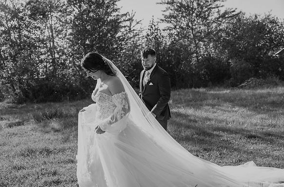 Bride walking in wedding dress with groom looking on grassy field.