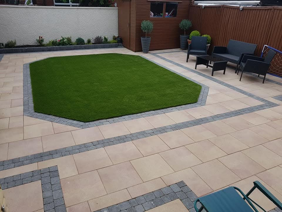 The image shows a modern backyard patio featuring a central octagonal patch of artificial turf bordered by dark grey cobblestone edging and light beige or cream paving slabs.