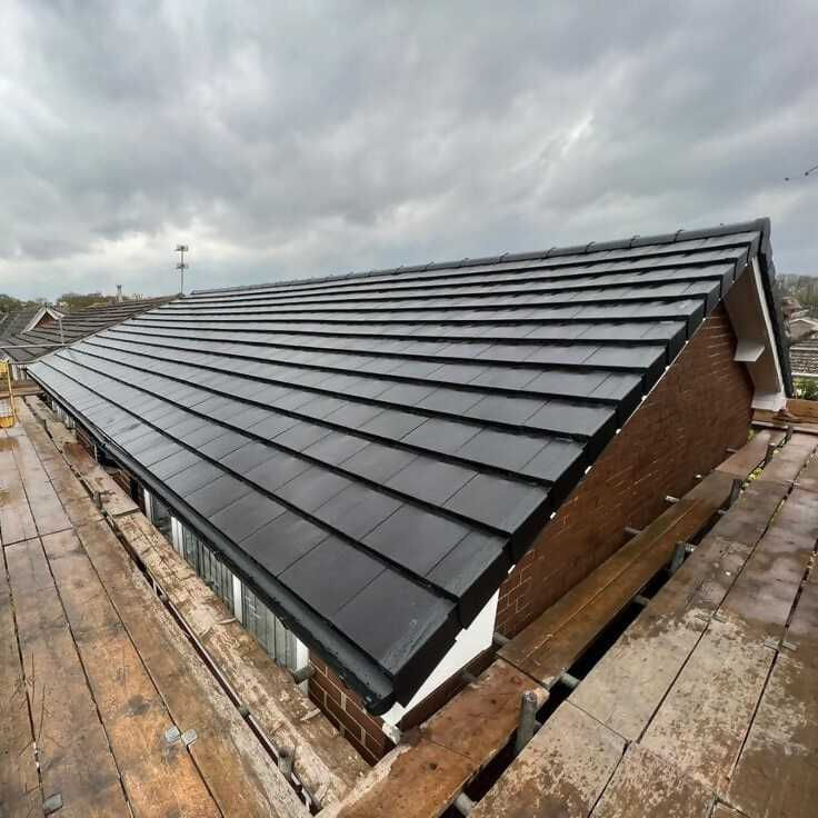 A view of a sloped roof covered with dark shingles, with wooden scaffolding visible in the foreground and a cloudy sky in the background.