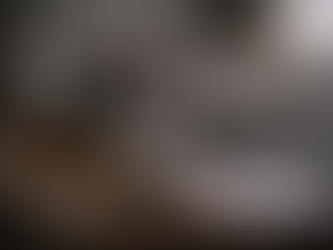 Dimly lit bedroom scene with a wooden bedside table holding a whiskey bottle, half-filled glass, cigarettes, car keys, tissues, and paperwork, with an unmade bed blurred in the background, suggesting distress and domestic tension.