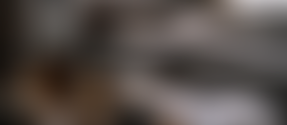 Dimly lit bedroom scene with a wooden bedside table holding a whiskey bottle, half-filled glass, cigarettes, car keys, tissues, and paperwork, with an unmade bed blurred in the background, suggesting distress and domestic tension.