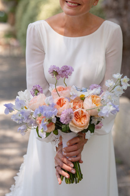 Close-up of the bride holding her wedding bouquet, with soft seasonal flowers in shades of lilac, white, and beige, complementing her dress beautifully.