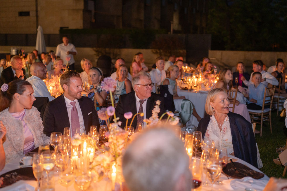 Guests seated at beautifully decorated tables with glowing candles, listening to a speech during a nighttime wedding dinner under a magical ambiance.