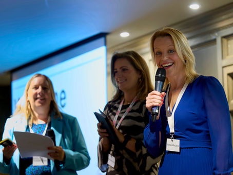 Mehalah is talking on stage at an event with two guests. Mehalah holds a microphone and smiles at the camera.