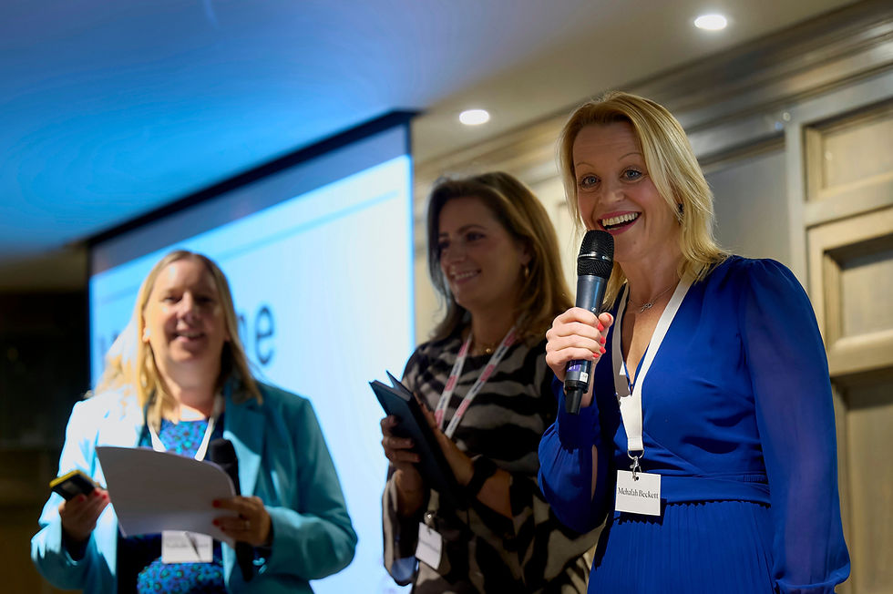 Mehalah is talking on stage at an event with two guests. Mehalah holds a microphone and smiles at the camera.
