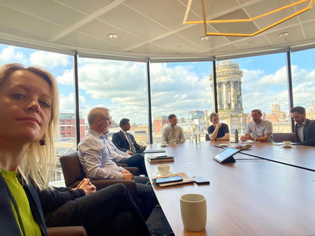 Mehalah Beckett looking at the camera, while sat at a boardroom table. There are people sat at the table discussing something and the window looks out onto a city skyline.