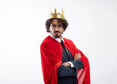 Free photo serious young caucasian superhero man in optical glasses wearing suit with red cloak and crown stands sideways with crossed arms