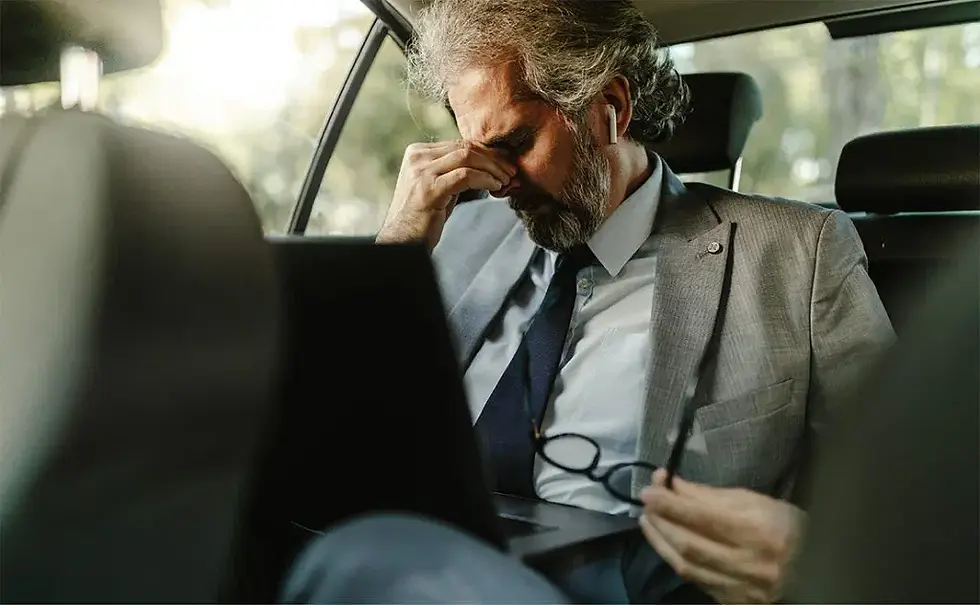 Uomo over 40 in auto, con abito elegante, affaticato e stressato, che si tocca il volto mentre tiene gli occhiali in mano.