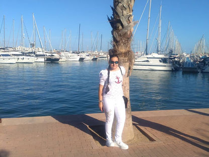 Carmen Galvañ de blanco y gafas de sol posa junto a una palmera en un puerto deportivo, con yates amarrados y cielo azul.