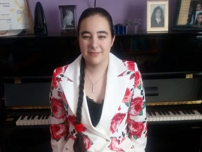 Carmen Galvañ con blazer blanco de flores y trenza frente a un piano negro, con fotos y un certificado encima.