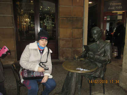 Carmen Galvañ sentada junto a una estatua de bronce leyendo en una cafetería; al fondo se lee Peletetos desde 1939.