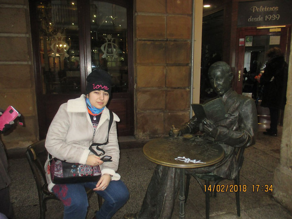 Carmen Galvañ sentada junto a una estatua de bronce leyendo en una cafetería; al fondo se lee Peletetos desde 1939.
