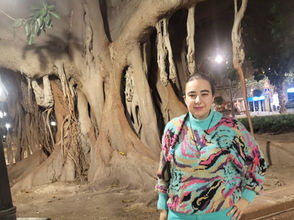 Carmen Galvañ junto a un gran árbol de raíces aéreas, con suéter multicolor y pantalón turquesa en una plaza urbana.