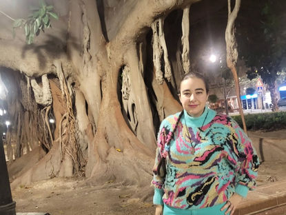 Carmen Galvañ junto a un gran árbol de raíces aéreas, con suéter multicolor y pantalón turquesa en una plaza urbana.