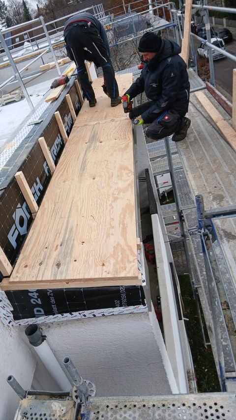 Zwei Dachdecker montieren Holzplatten am Rand eines Flachdachs während der Sanierung