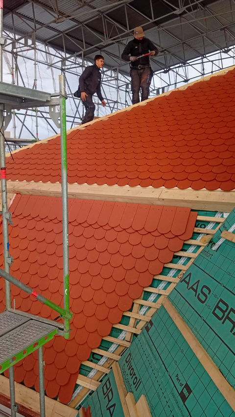 Zwei Dachdecker verlegen rote Dachziegel auf einem geneigten, eingerüsteten Dach. (Dach Mergler Dachdecker München)