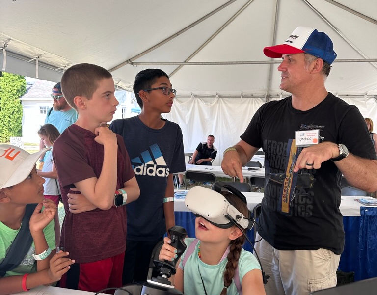 Girl in VR headset with controllers, watched by boys and man.