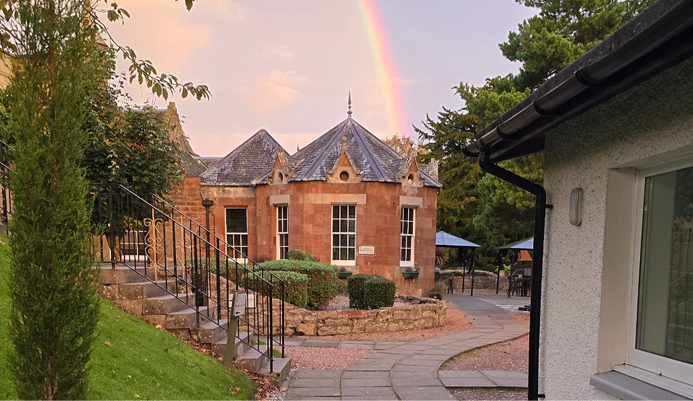 Stone building with pointed roof features a rainbow in the sky. Surrounded by greenery, steps, and a path. Calm and picturesque setting.