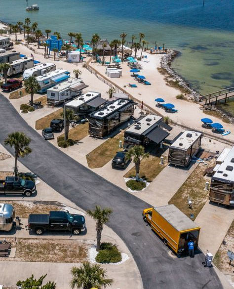 Aerial view of RVs parked near a beach with palm trees and blue umbrellas. A yellow truck is parked on the road. The water is calm.