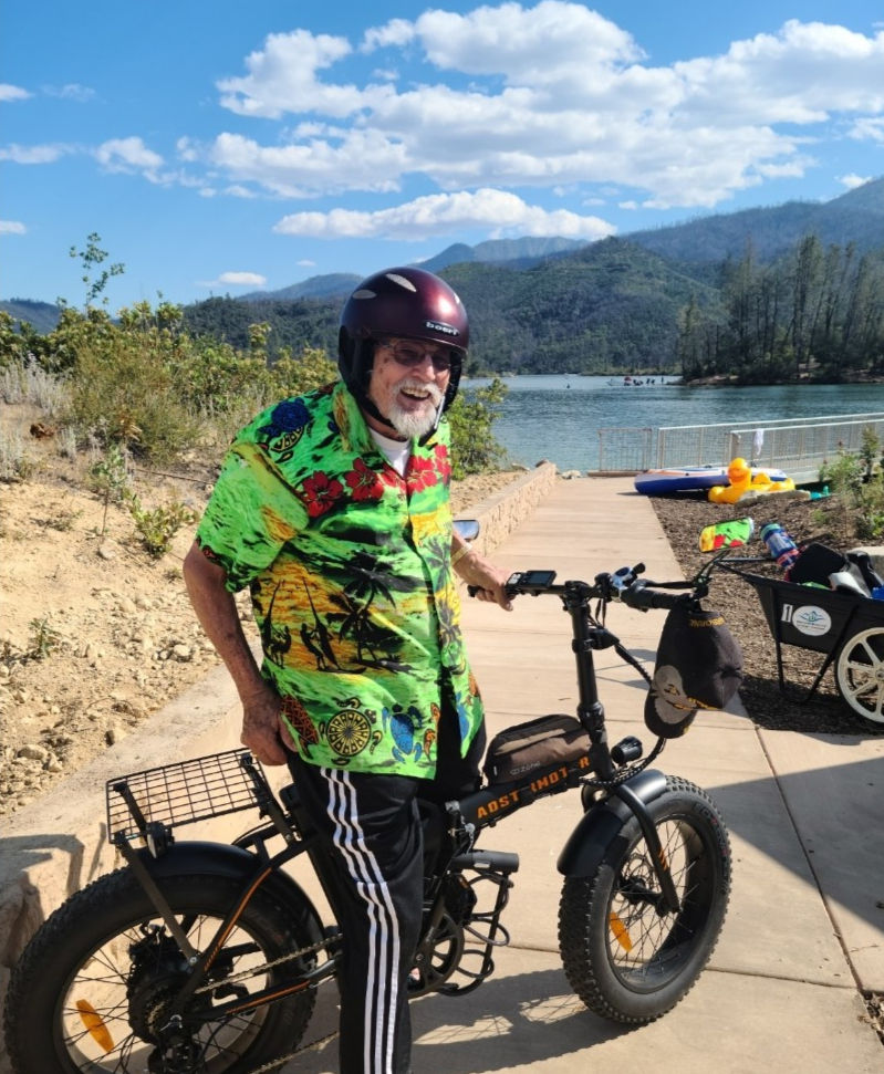 Elderly man in a colorful shirt and helmet smiles next to an electric bike by a lake with mountains and blue sky in the background.