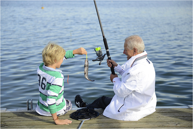 A child and elderly man sit on a dock fishing by a lake. The boy holds a fish, and the man has a fishing rod. Both appear happy.