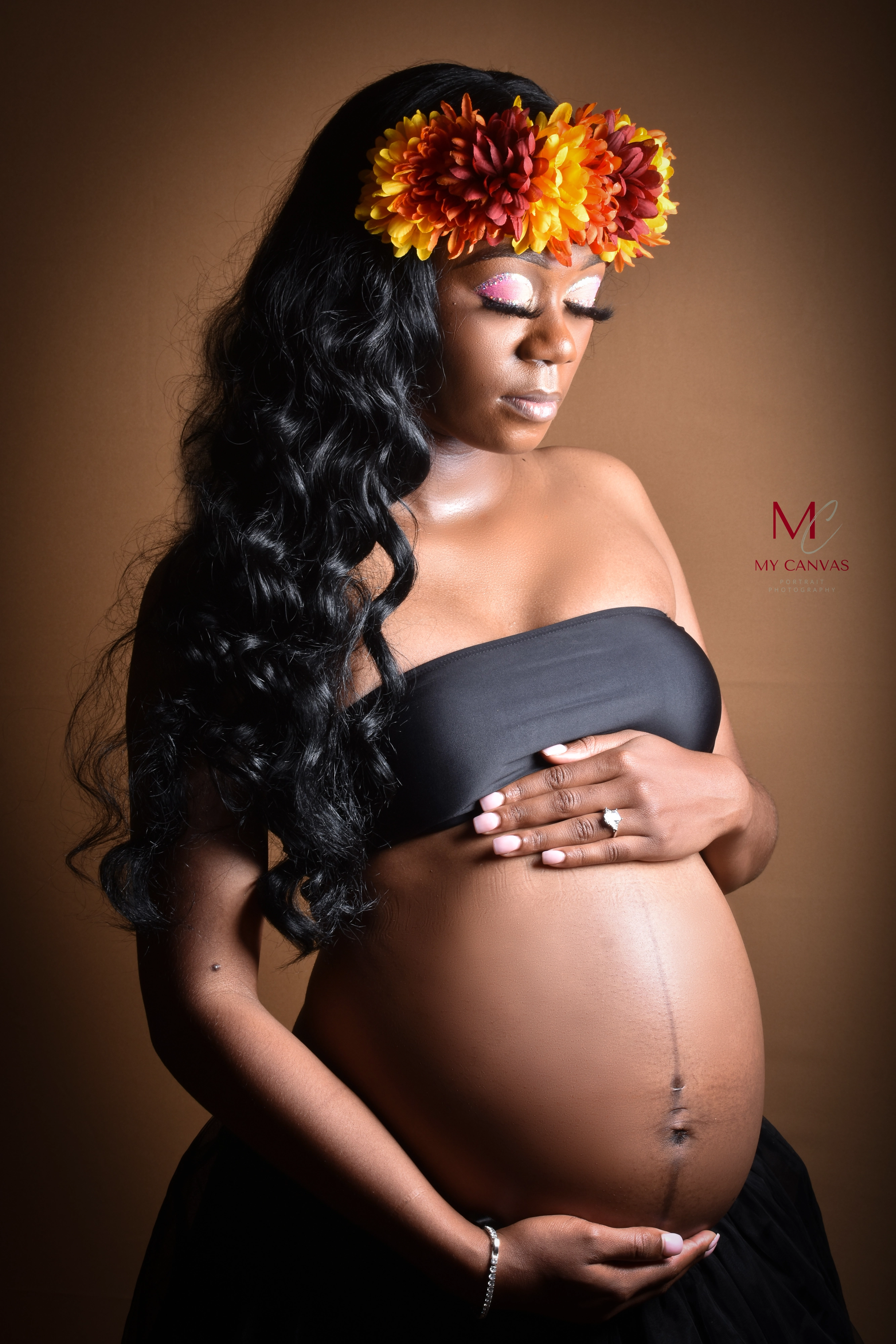 A pregnant woman in Killeen gazes downward affectionately at her belly, framed by a crown of vibrant orange flowers, creating a regal and nurturing image.