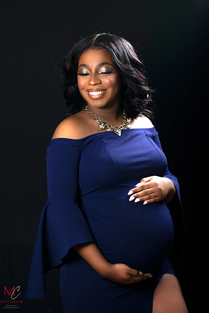 A joyful pregnant woman in Killeen stands solo, her smile bright and contagious, dressed in an elegant off-shoulder navy dress that hugs her form and highlights her baby bump.