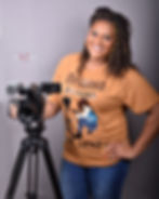 Smiling woman in orange shirt with "Melanin Behind The Lens" text, standing by a camera on tripod, gray background.