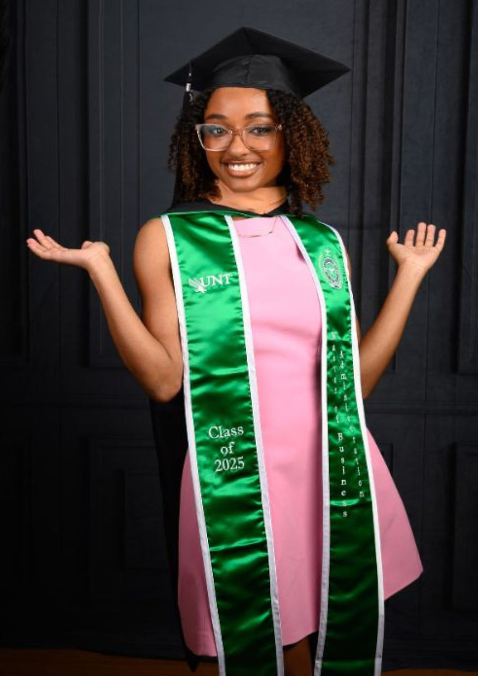 Graduate wearing a black cap, green graduation stole, and pink dress during a full-length portrait session.