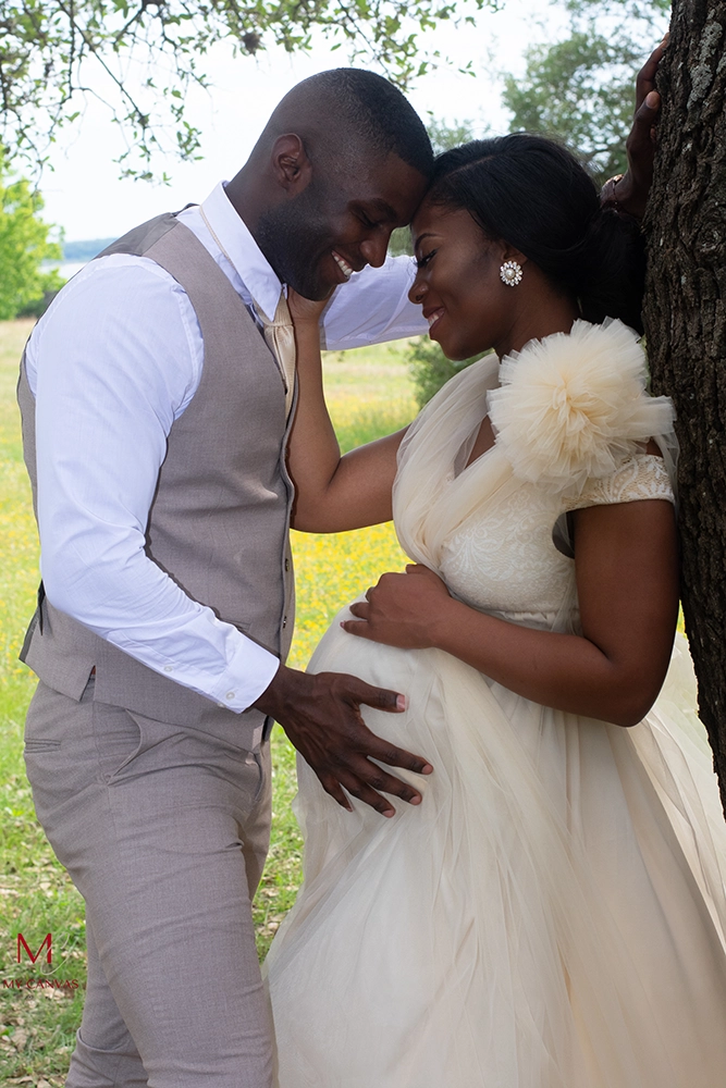 A couple in Killeen, Texas, intimately connects in a sunlit setting, the pregnant woman in white, as they both tenderly focus on their future child.