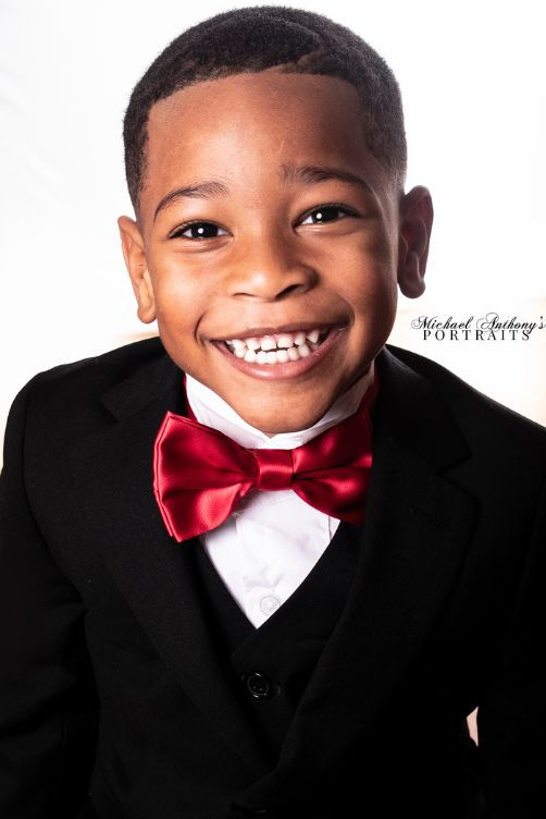 Young boy smiling in a black suit with a red bow tie during a studio portrait session