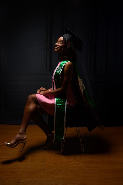 This studio portrait shows a graduate seated in profile, wearing a pink dress and a green stole. The lighting creates a dramatic, elegant side silhouette that highlights her poise and confidence. The dark backdrop and wooden flooring add a timeless, classic feel to the image. Her smile suggests pride and excitement for the future ahead.