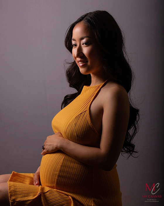 A serene moment is captured in Killeen as an expectant mother cradles her bare baby bump while nestled in a cozy, knitted yellow dress, her profile silhouetted against a soft grey backdrop.
