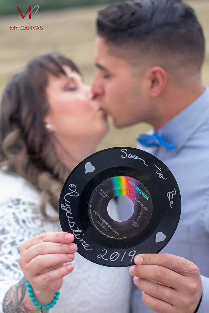 Couple kissing, holding a vinyl record with "Soon to Be Augustine 2019" written on it. Soft focus background, romantic mood.