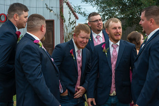 A candid moment among groomsmen, dressed sharply in matching suits with pink ties, sharing a light-hearted conversation that reflects the strong bonds of friendship.