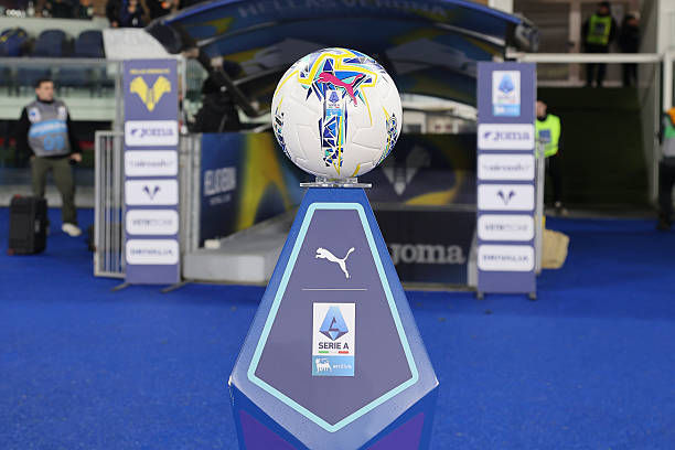 Football on a stand with Serie A logo, blue floor, and Puma branding. Background shows stadium entry with security and signage.