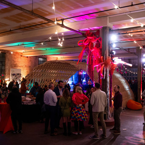 A social gathering at an art show. A large wooden sculpture is seen in the background among the groups of people.