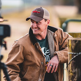 Logan Duvall smiling and leaning against a wooden fence, wearing a brown jacket, green shirt, and a cap with a red truck design.