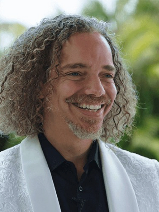 Dr. Henry Ealy smiling outdoors in a white patterned jacket with a dark shirt, standing in front of green foliage.