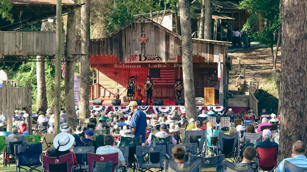 A large outdoor concert at Salmon Lake Park in Grapeland, Texas. A crowd of people sits in folding chairs and camping chairs on the grass under tall pine trees, facing a wooden stage with a red backdrop and a large Texas star. Musicians with guitars and a double bass perform on stage while audience members enjoy the live music on a sunny day. American and Texas flags are displayed on the stage.