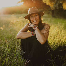 Jacqueline Bourne sitting in a sunlit field, smiling warmly while wearing a tan hat, black dress, and colorful bracelets, with trees glowing in the background.