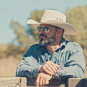 AJ Richards, founder of From The Farm, wearing a cowboy hat and sunglasses, leaning on a fence outdoors.