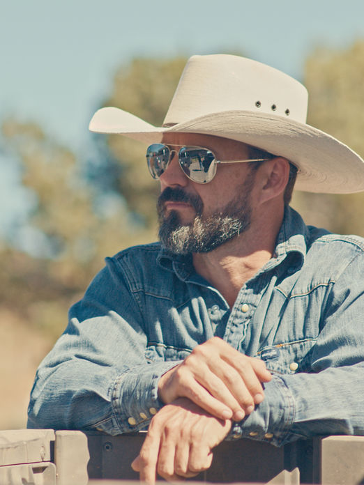 AJ Richards, founder of From The Farm, wearing a cowboy hat and sunglasses, leaning on a fence outdoors.