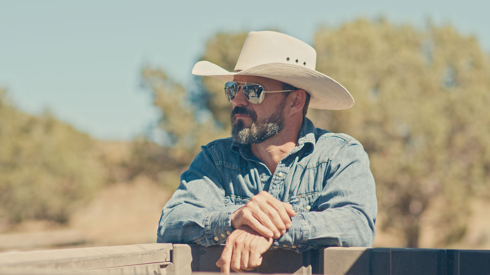 AJ Richards, founder of From The Farm, wearing a cowboy hat and sunglasses, leaning on a fence outdoors.