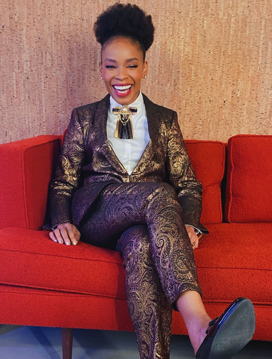 Amber Ruffin rocking an Afro puff, laughing, and sitting on a red couch wearing a gold and burgundy paisley suit.