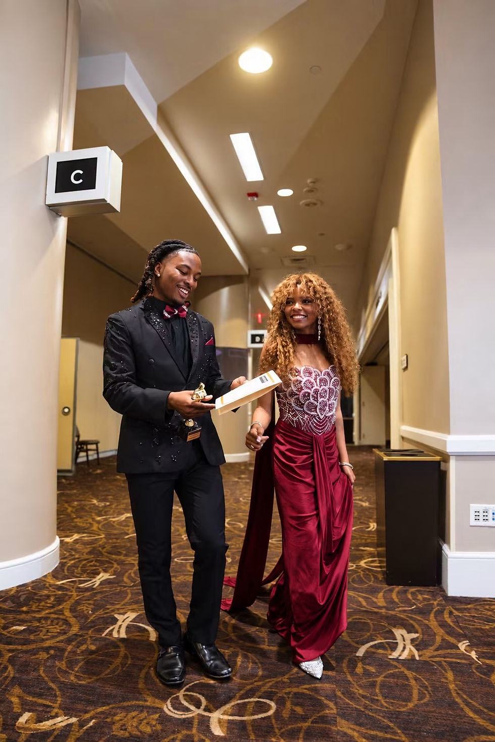 Central State Royal Court pair walking through an indoor corridor in formal attire; one carries awards.