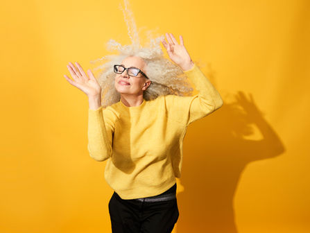 A senior woman in a yellow sweater and dark glasses dancing in the sun with a bright yellow background.