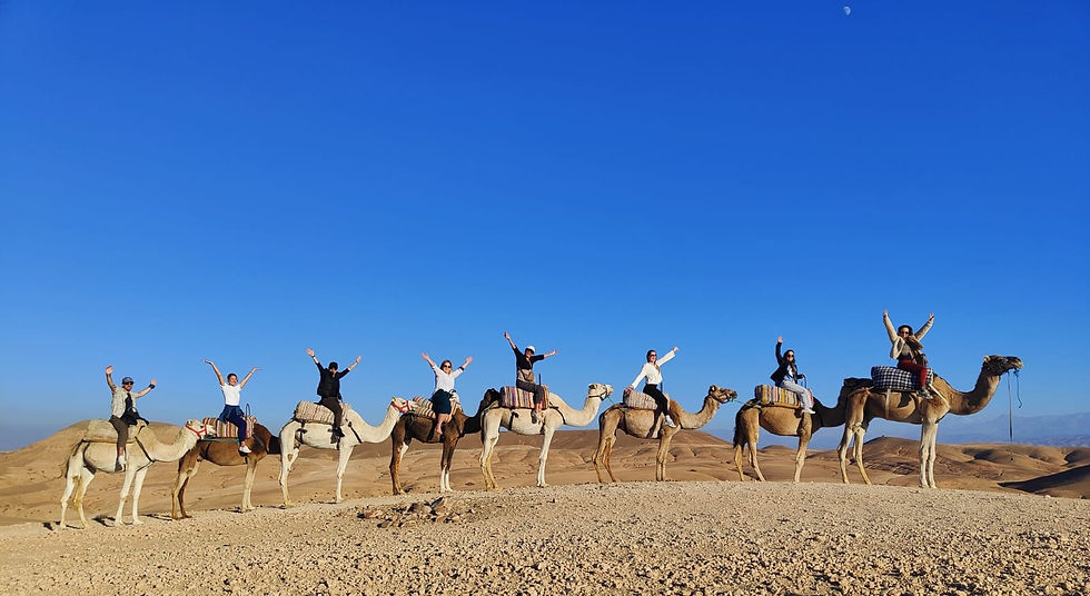 People ride camels in a desert, with arms raised, under a clear blue sky. The mood is joyful, with barren hills in the background.