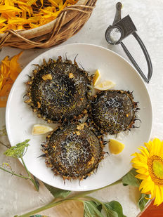 Grilled sunflower heads with cheese and lemon on a white plate. Sunflower petals in a basket, yellow flower, and grater in the background.