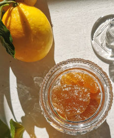 Sunlit lemon beside a crystal jar of candied citrus peels on a white surface, with its crystal lid at right.